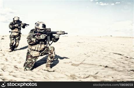 Marine rider in helmet and camo uniform, standing on one knee, aiming service rifle, supporting with fire running behind teammate. United States army special forces soldiers attacking enemy in desert. Marine riders moving with caution through sands