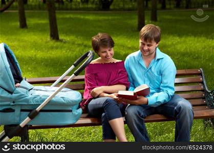 Married couple on walk with the baby sit on a bench in park and read the book on a background of a lawn. Family walk