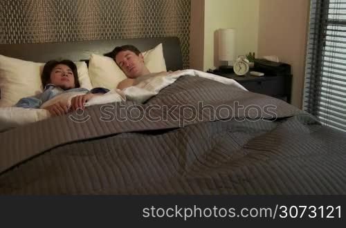 Married people, marriage relationship, husband and Asian wife at home. Early morning with tired man snoozing alarm clock, waking up and Japanese woman staying in bed. Interracial couple lifestyle