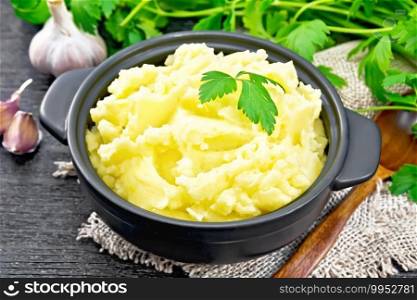 Mashed potatoes in a black saucepan and a spoon on a burlap napkin, garlic, parsley on wooden board background