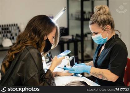 Masked beautician, painting her client&rsquo;s nails in a beauty center, while she records a video with her smartphone.. Masked beautician, painting her client&rsquo;s nails while she records a video with her smartphone.