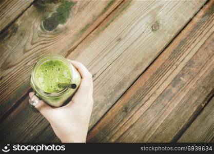 Mason jar mug filled with green smoothie In hand on wooden rustic table. Green healthy food concept