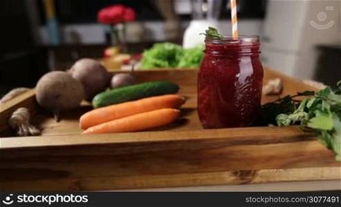 Mason jar of red fresh beet smoothie with striped straw and mint leaves for decoration with vegetables in wooden box tray over kitchen background closeup. Healthy eating and dieting concept.