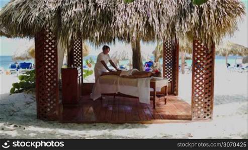 Massage therapist providing spa treatment for a woman in gazebo with straw roof on the beach