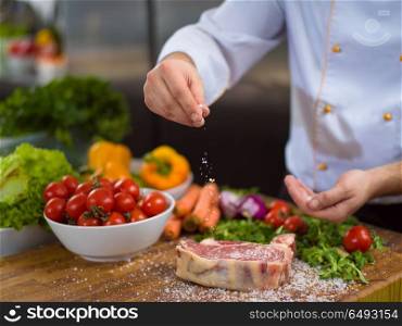 Master Chef hands putting salt on juicy slice of raw steak with vegetables around on a wooden table. Chef putting salt on juicy slice of raw steak