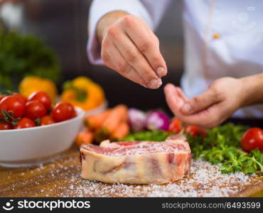 Master Chef hands putting salt on juicy slice of raw steak with vegetables around on a wooden table. Chef putting salt on juicy slice of raw steak