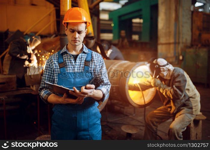 Master with notebook and welder in mask on factory, man works with metal pipe on factory, welding skill. Metalworking industry, industrial manufacturing of steel products. Master with notebook and welder on factory