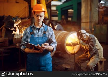Master with notebook and welder in mask on factory, man works with metal pipe on factory, welding skill. Metalworking industry, industrial manufacturing of steel products. Master with notebook and welder on factory