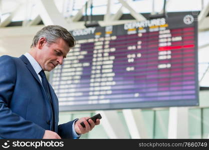 Mature attractive businessman using smartphone while standing against flight display screen in airport