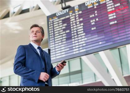 Mature attractive businessman using smartphone while standing against flight display screen in airport