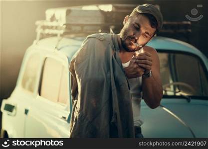 Mature bearded man lighting a cigarette, a retro car with suitcases on a roof rack on background