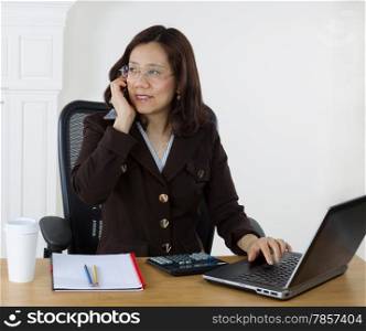 Mature business woman talking on cell phone while working with laptop, calculator and notepad on desktop. White wall background.