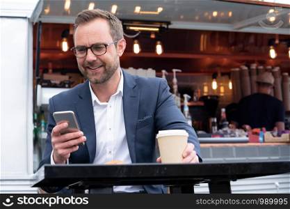 Mature Businessman Checking Mobile Phone Sitting At Outdoor Coffee Shop