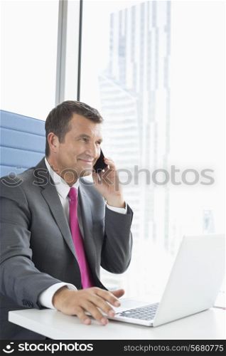 Mature businessman talking on cell phone while using laptop in office