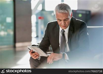 Mature businessman using digital tablet while checking time on his watch in airport