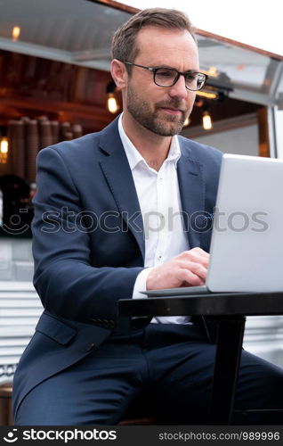 Mature Businessman Working On Laptop By Outdoor Coffee Shop