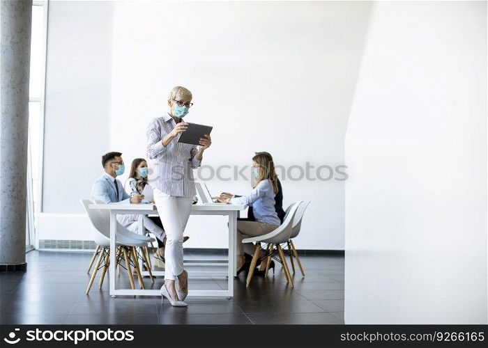 Mature businesswoman with protective mask using digital tablet in office in front of her team