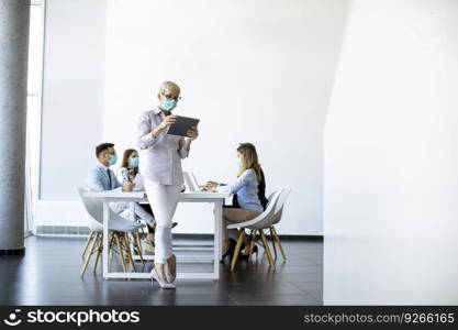 Mature businesswoman with protective mask using digital tablet in office in front of her team