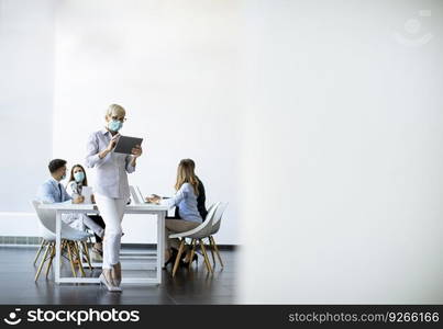 Mature businesswoman with protective mask using digital tablet in office in front of her team