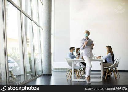 Mature businesswoman with protective mask using digital tablet in office in front of her team