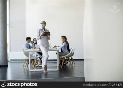 Mature businesswoman with protective mask using digital tablet in office in front of her team