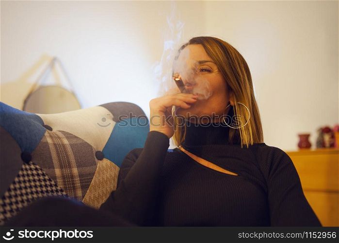 Mature caucasian woman in black smoking cigarillo at home while sitting in a colorful chair in the evening