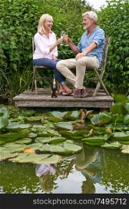 Mature Couple Celebrating With Champagne Sitting On Chairs On Wooden Jetty By Lake