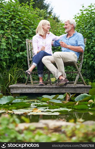 Mature Couple Celebrating With Champagne Sitting On Chairs On Wooden Jetty By Lake