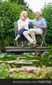 Mature Couple Celebrating With Champagne Sitting On Chairs On Wooden Jetty By Lake