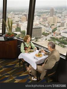 Mature couple dining in fancy restaurant by window with rooftop view of urban landscape.