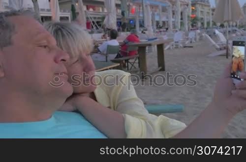 Mature couple is cuddling in beach lounger and taking selfie shot at smartphone.