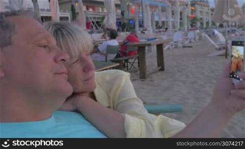 Mature couple is cuddling in beach lounger and taking selfie shot at smartphone.
