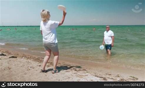 Mature couple is playing racket ball on the beach in sunny day, woman is standing on the sand, man is in water.