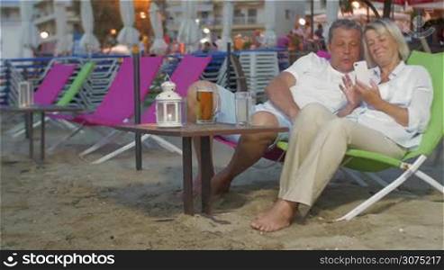 Mature couple is resting on the beach on sun lounger, they are watching something in smartphone and smiling