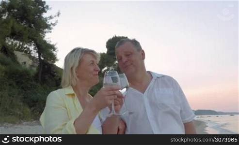 Mature couple is standing by the sea in twilight. They are holding wine glasses in hands, clanging them and drinking.