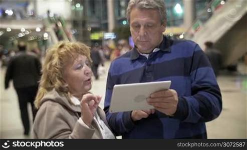Mature couple is standing in some public place, man is holding tablet PC in hands, woman is explaining something emotionally.