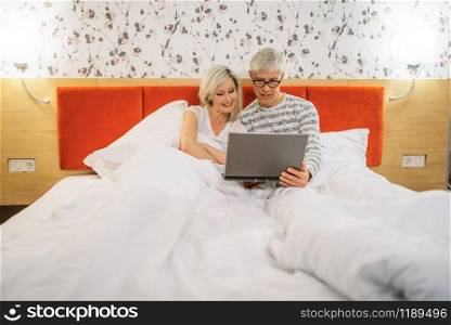 Mature couple looking on laptop in bedroom before sleeping. Adult man in glasses and woman lying in bed