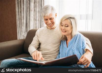 Mature couple sitting on couch and looking at old photo album, happy family. Adult husband and wife resting at home
