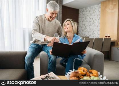 Mature couple sitting on couch and looking at old photo album, happy family. Adult husband and wife resting at home. Mature couple looking at old photo album