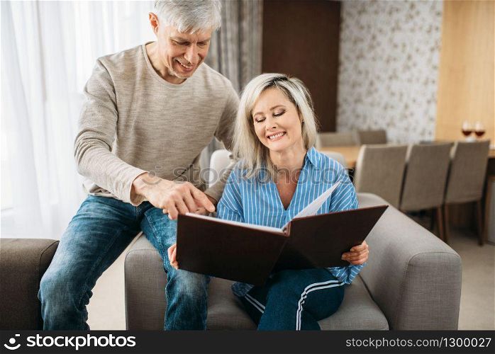 Mature couple sitting on couch and looking at old photo album, happy family. Adult husband and wife resting at home. Mature couple looking at old photo album