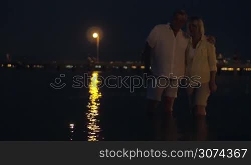 Mature couple standing in sea by the shore and looking at small candles floating on the water, golden light from lantern reflecting on the surface. Candles symbolizing their love