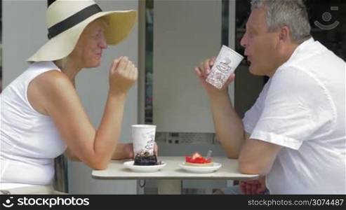 Mature family couple enjoying coffee and desserts in outdoor cafe. Woman in summer hat eating delicious chocolate cake and man having strawberry one