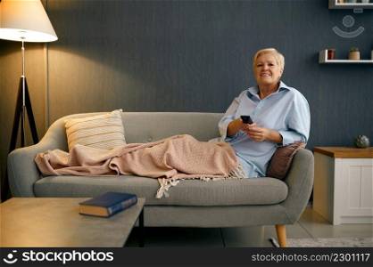 Mature grey-haired woman warming blanket and watching TV at home. Cozy evening on retirement. Mature woman wrapped in blanket watching TV