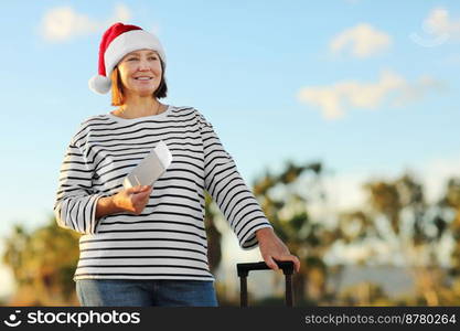 Mature happy woman in Santa hat, striped shirt is holding passport and ticket for trip on palms trees and sky background, New Year,Christmas winter holidays, tourism travel vacation concept on resort.. Mature happy woman in Santa hat, striped shirt is holding passport and ticket for trip on palms trees and sky background, New Year, Christmas winter holidays, tourism travel vacation concept on resort
