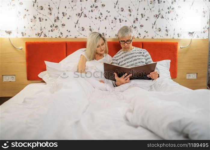 Mature husband and wife looks at photo album in bedroom. Adult man and woman lying in bed before sleeping, happy love couple