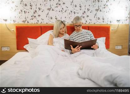 Mature husband and wife looks at photo album in bedroom. Adult man and woman lying in bed before sleeping, happy love couple