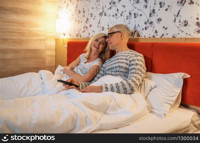 Mature love couple watching TV in bedroom before sleeping. Adult man in glasses and woman lying in bed. Mature love couple watching TV in bedroom