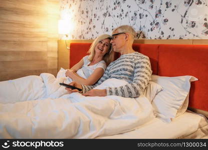 Mature love couple watching TV in bedroom before sleeping. Adult man in glasses and woman lying in bed. Mature love couple watching TV in bedroom