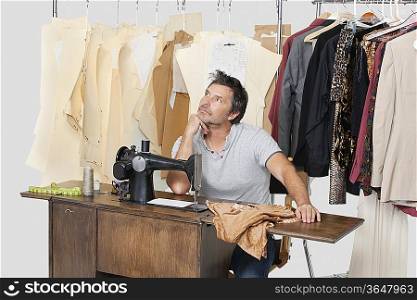 Mature male tailor thinking while sitting at sewing machine with fabric