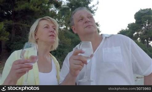 Mature man and woman are standing outdoor, drinking water from the glasses and talking.